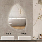 An organic teardrop-shaped wall mirror with an integrated LED light, mounted on a textured gray bathroom wall above a double sink vanity.