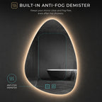 A teardrop-shaped bathroom mirror with warm LED lighting around its edge, shown on a dark background with text reading 'BUILT-IN ANTI-FOG DEMISTER.' The mirror reflects a bathtub scene, and callout icons indicate anti-fog demister functionality.
